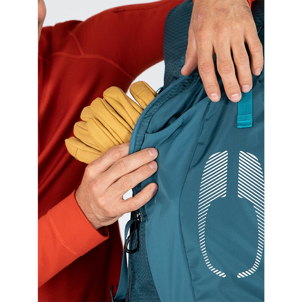 A close-up shows hands pulling a pair of yellow leather gloves out of the large, vertical zippered front panel pocket of a blue Osprey backpack