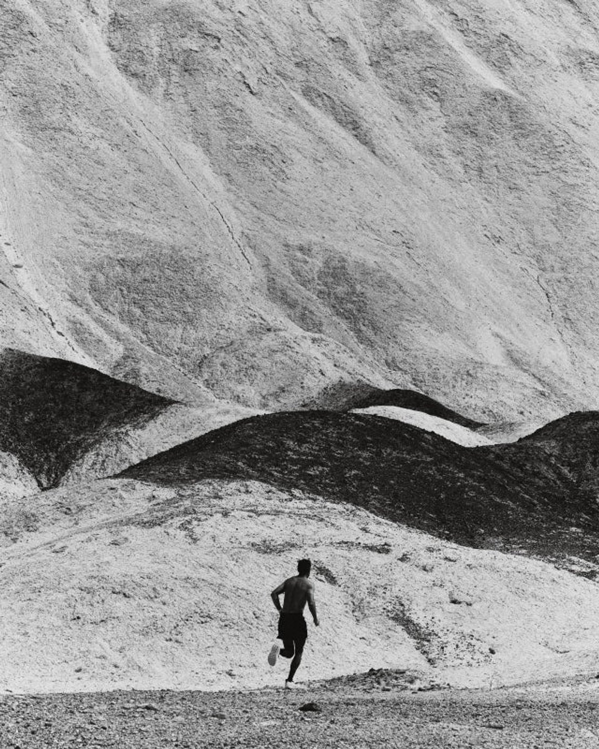 Black-and-white wide shot of a shirtless male runner as a small figure running toward layered pale desert hills.