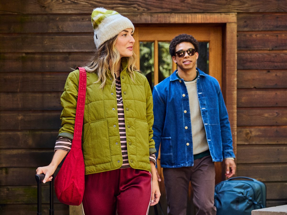 Two people leaving a wood cabin with luggage, wearing casual cold-weather layers including a quilted vest, beanie, and denim jacket.