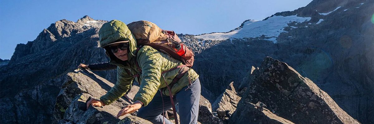 Female mountaineer rock climbing on a steep alpine ridge wearing yellow climbing helmet and green insulated jacket with backpack in bright daylight conditions.