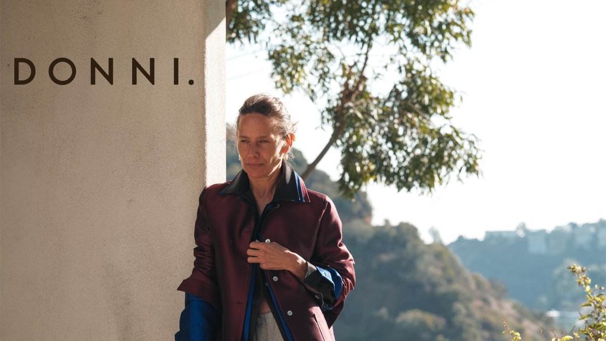 Woman wearing a burgundy DONNI. jacket standing against a white wall with scenic hillside landscape background.