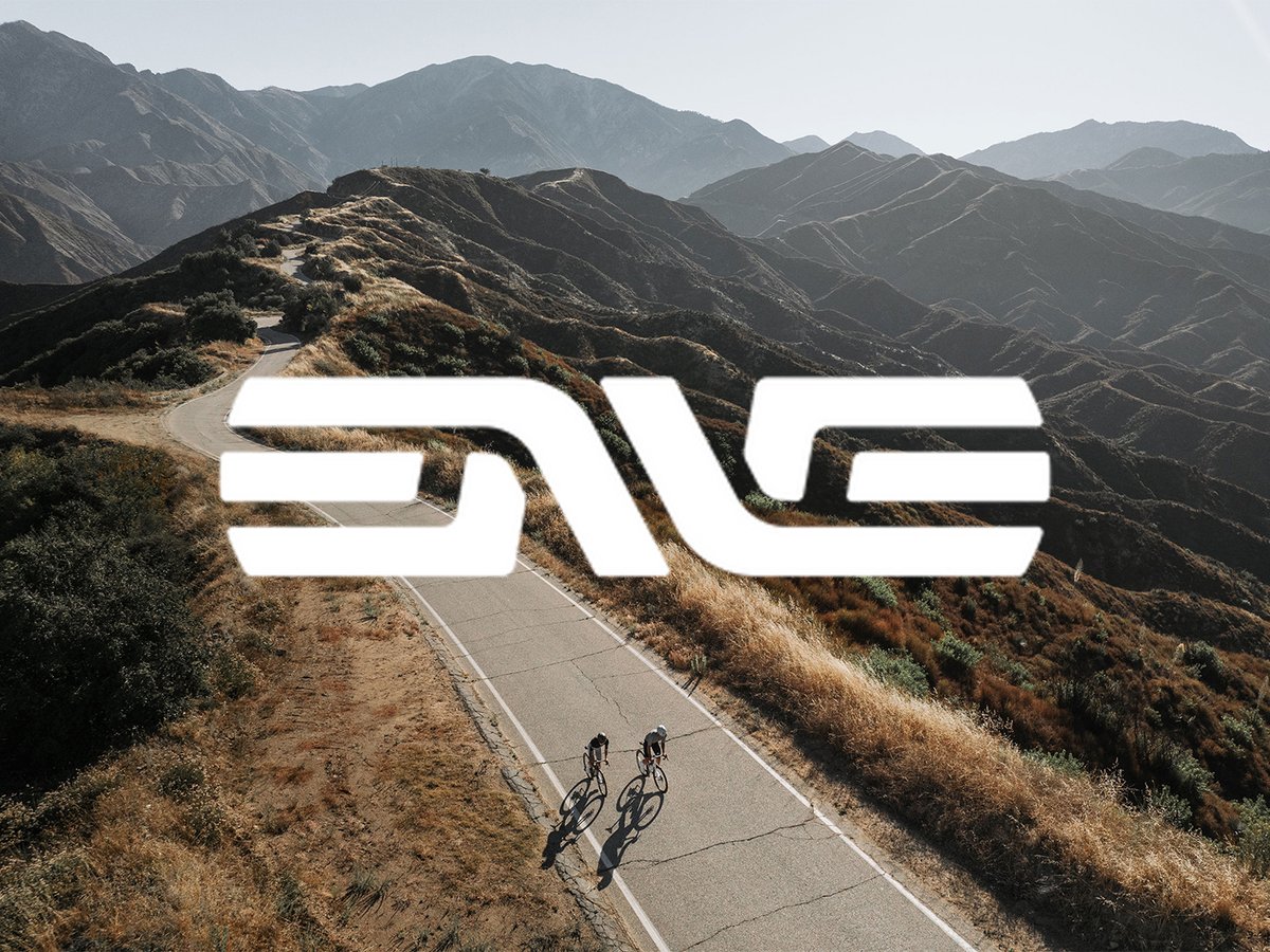 White envelope logo overlayed on mountain road with two cyclist in the foreground.
