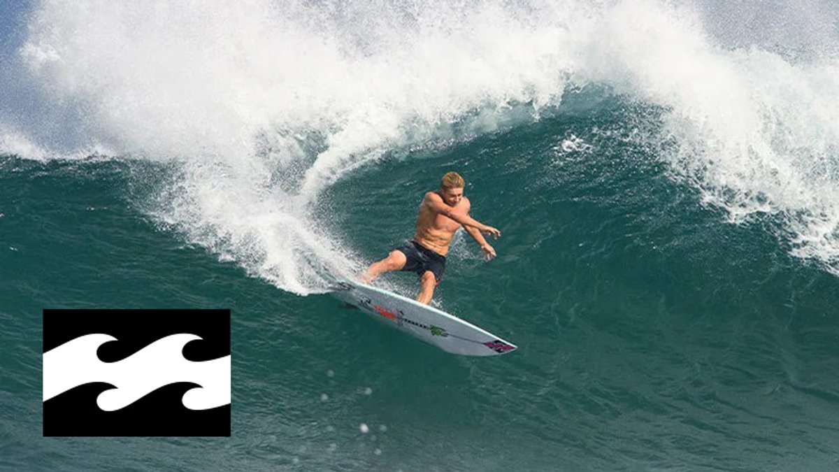 A shirtless male surfer rides a large wave on a surfboard while the Billabong logo is displayed in the lower left corner.