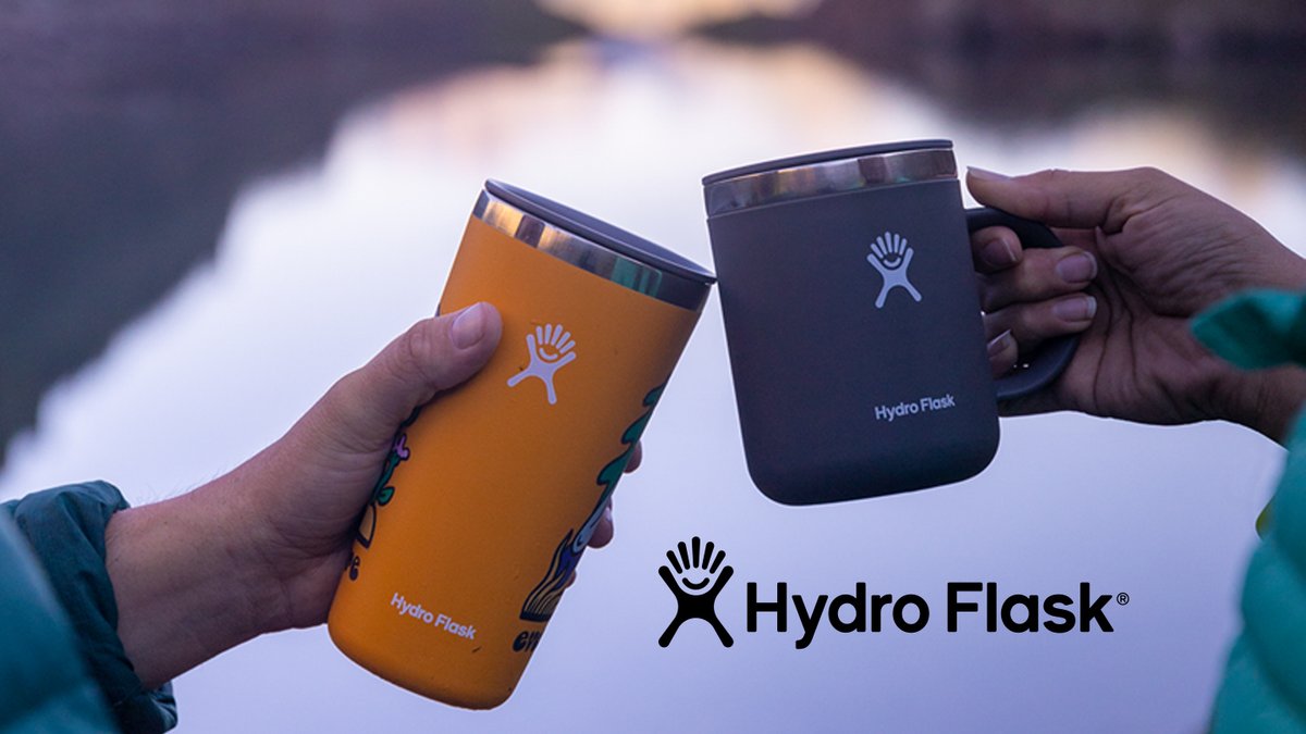 Two hands toast an orange Hydro Flask tumbler and a grey Hydro Flask mug against a blurred lakeside background at dusk.