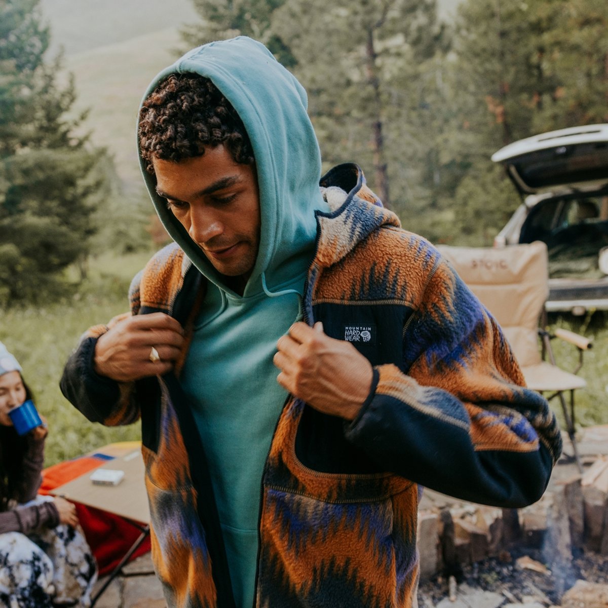 A man at a mountain campsite wearing a blue hoodie pulls on a flannel jacket.