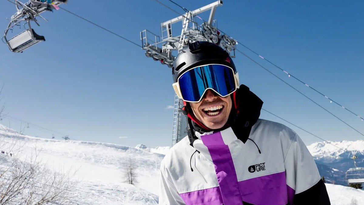 Happy skier wearing white and purple Picture brand jacket and blue ski goggles standing on snowy mountain slope with ski lift in background.