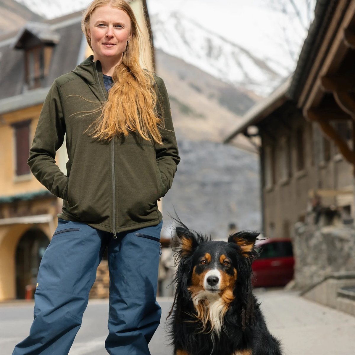 Woman with long reddish-blonde hair wearing an olive green zip-up hoodie and blue pants standing with a black and tan border collie dog on a mountain village street.