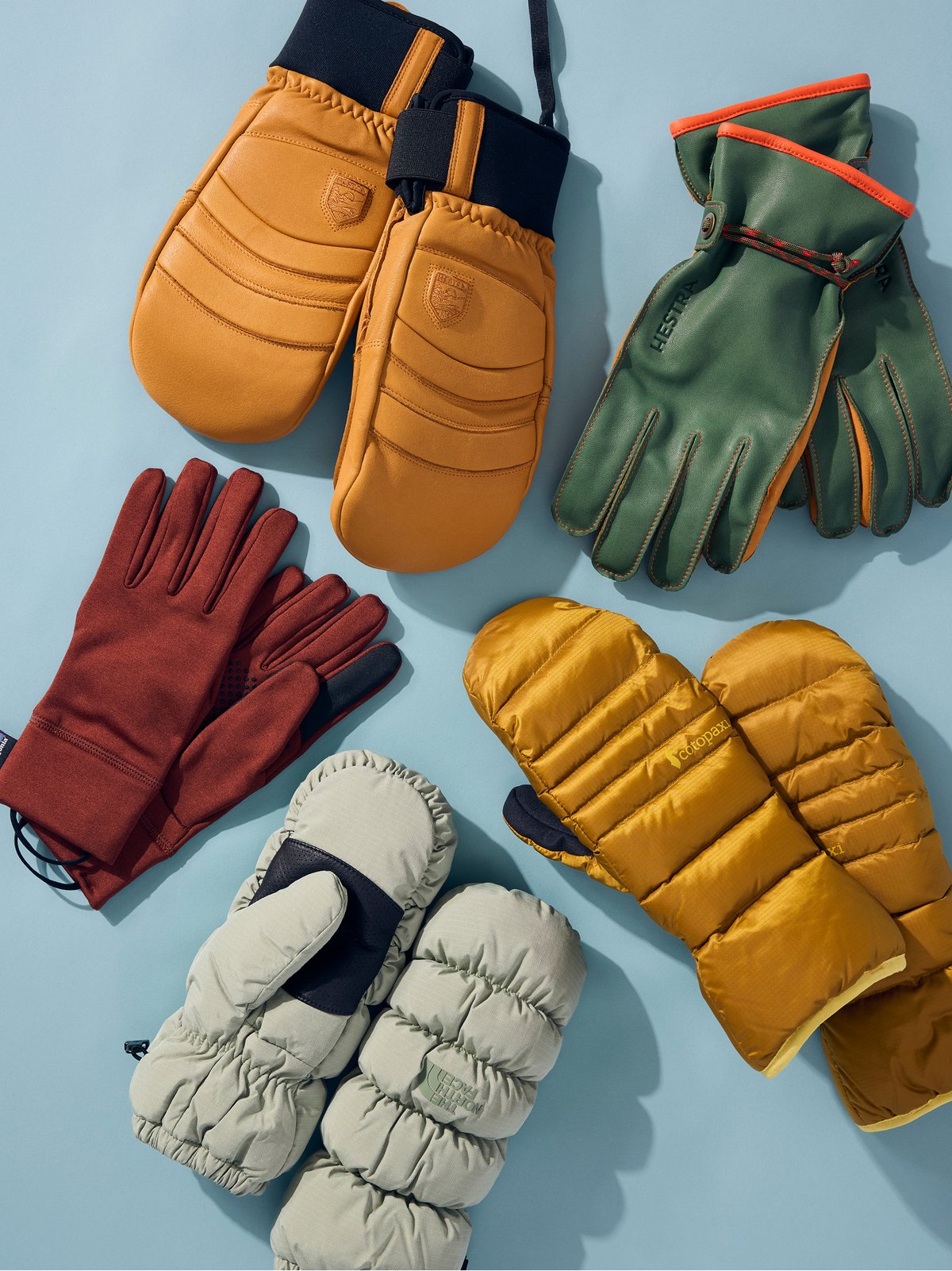A variety of puffy mittens and leather gloves arranged on a light blue background.