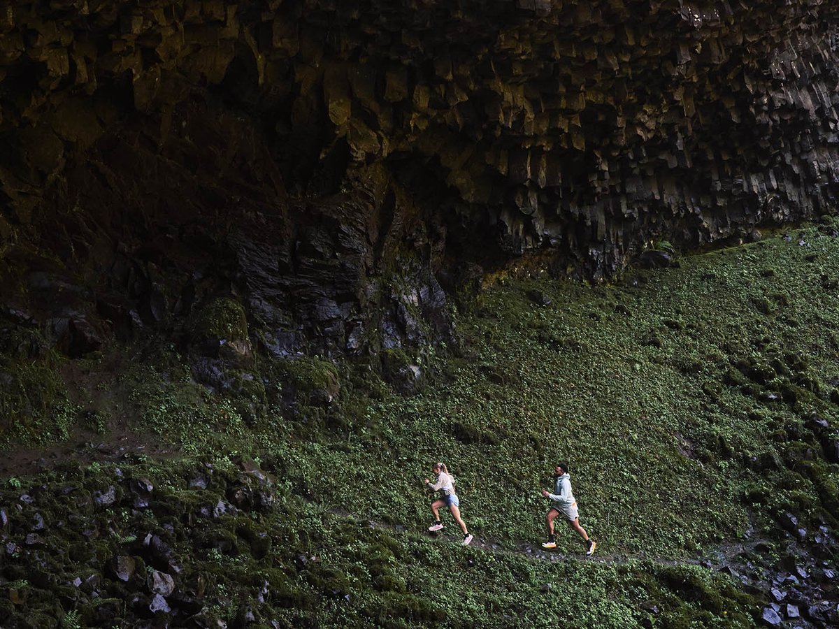 Two runners on trail in front of black rock cliff face.