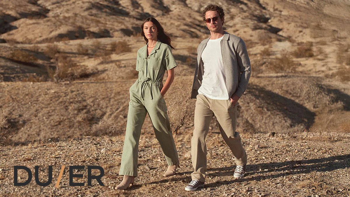 Two people wearing neutral-toned casual outdoor DUER apparel stand on a desert landscape, showcasing sustainable summer clothing from DUER's collection.