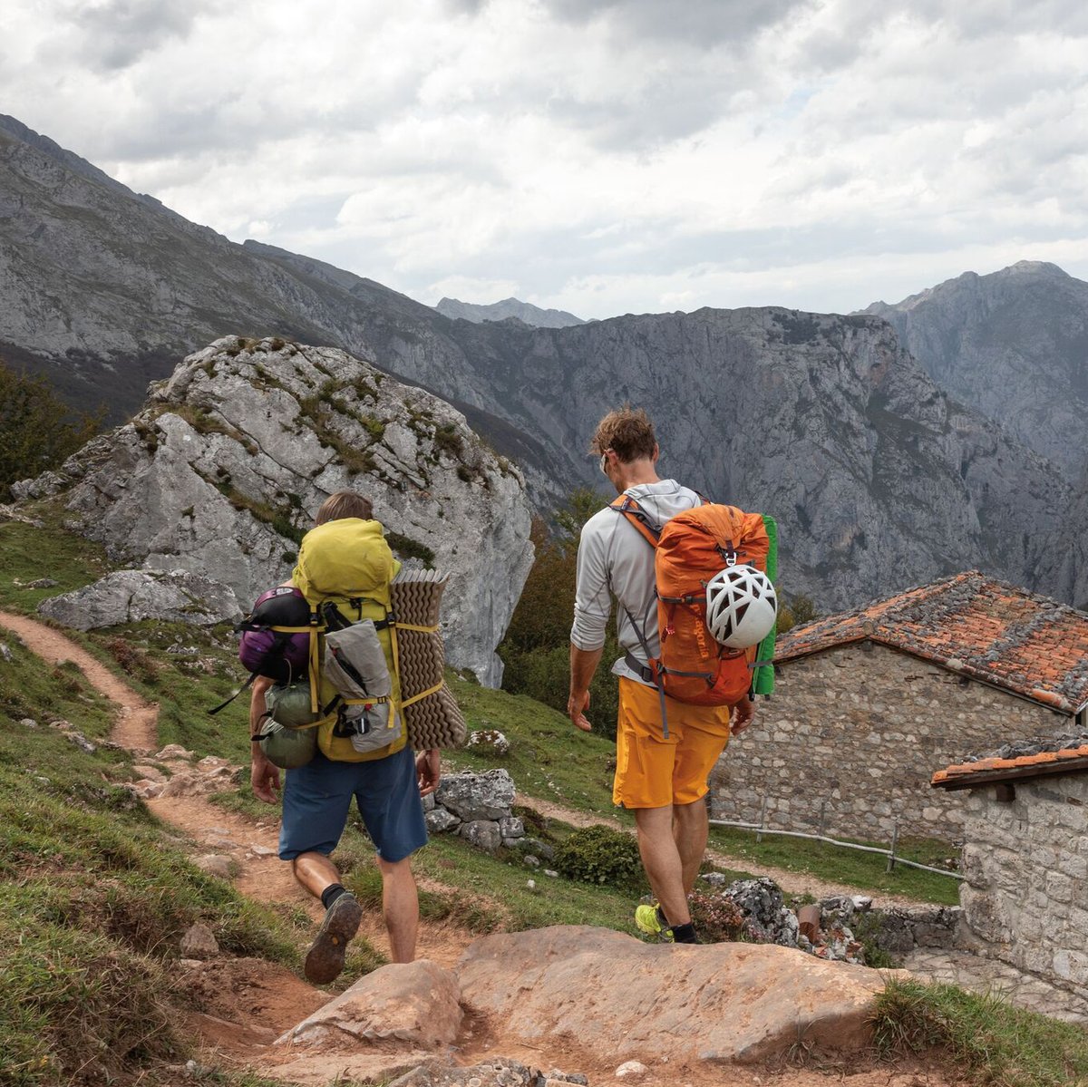 Two hikers with backpacks and helmets trekking on a mountain trail with dramatic rocky peaks and a stone building in an alpine landscape.
