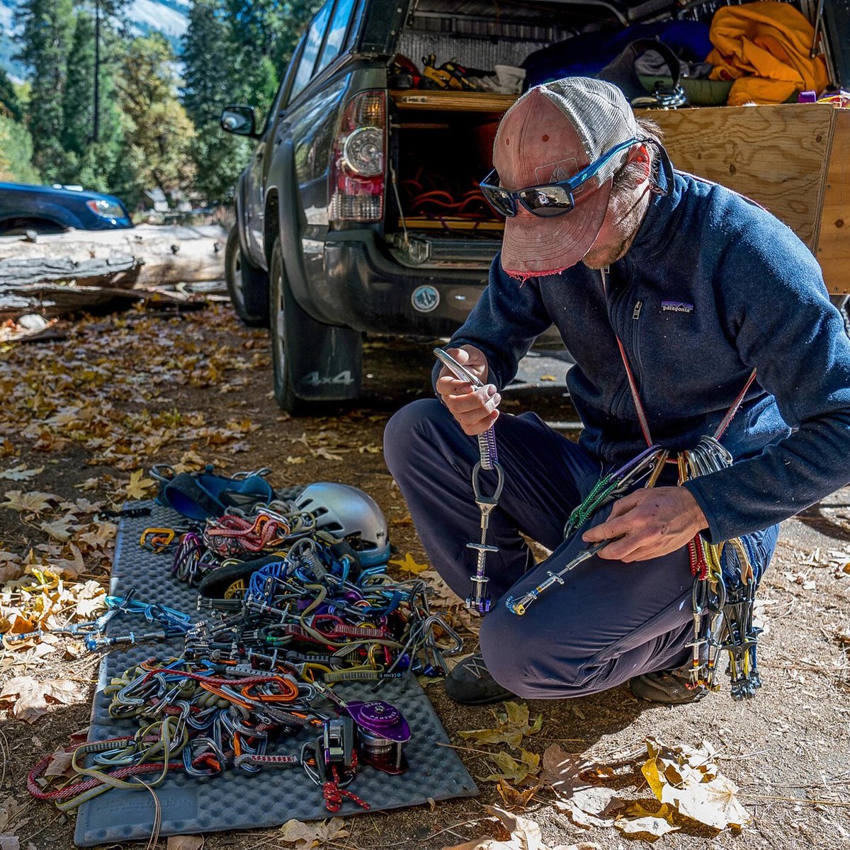 Experienced climber organizing colorful climbing ropes, carabiners, and safety gear laid out on ground near van in forest campsite.