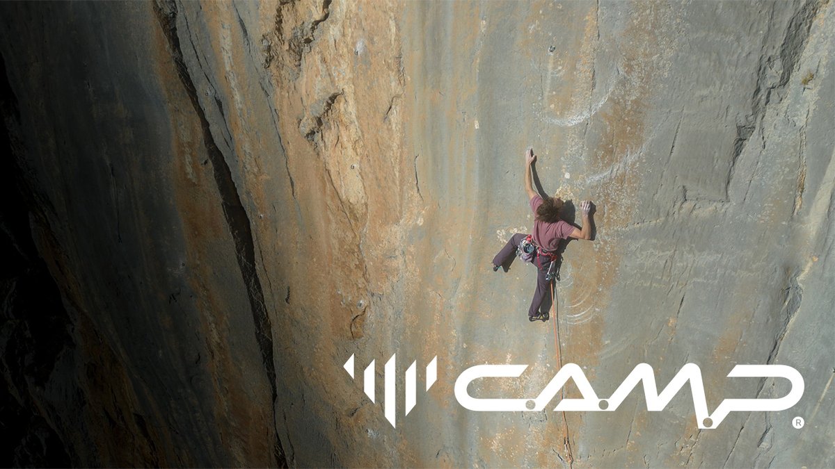 A rock climber in a red shirt scales a massive, sheer limestone cliff face above the CAMP brand logo.
