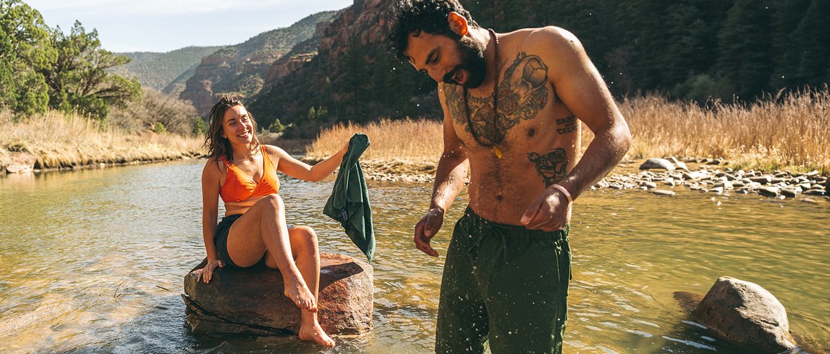 Person sitting on a rock in a river holding using Coalatree Microfiber Towel while friend stands in the water laughing. 
