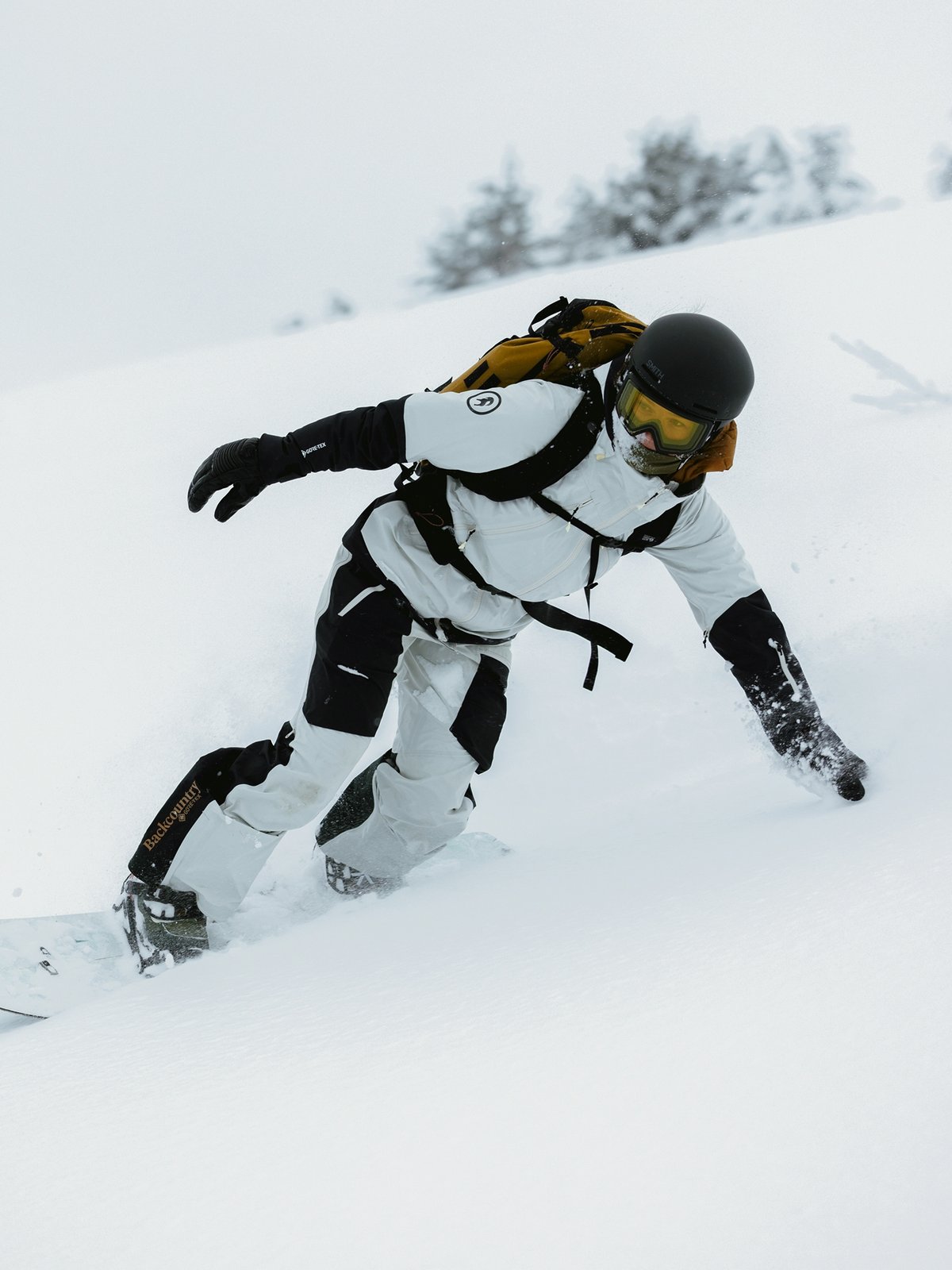 A snowboarder wearing a white jacket and pants, black helmet, yellow goggles, and a brown backpack carves through deep snow on a mountain slope.