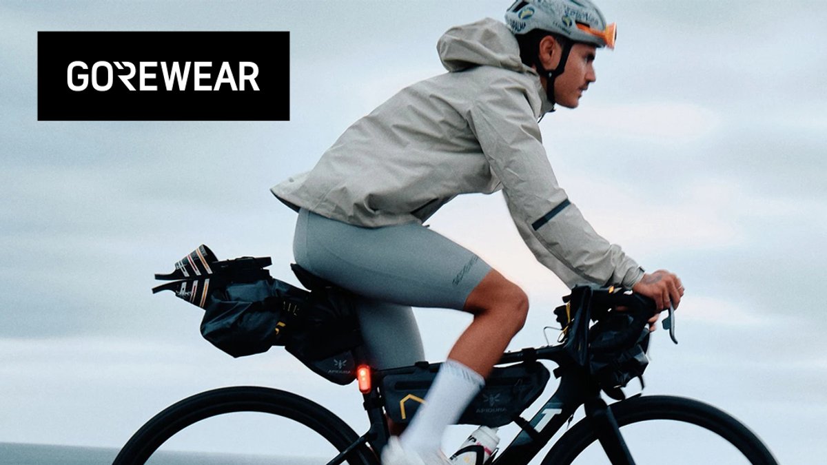A cyclist in a grey GOREWEAR jacket and matching shorts rides a bike loaded with bikepacking bags along a cloudy coast.