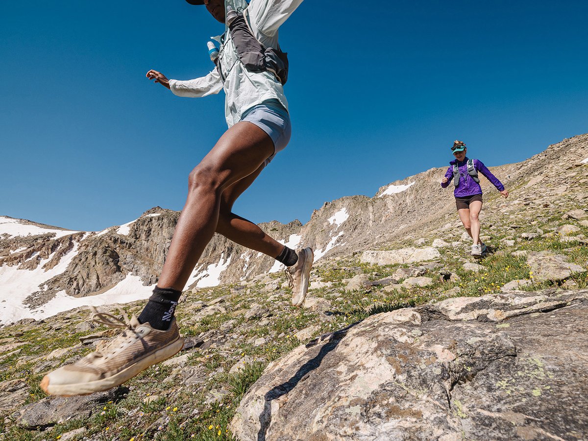 Two trail runners descending a rocky alpine meadow with snow-patched peaks under a deep blue sky.
