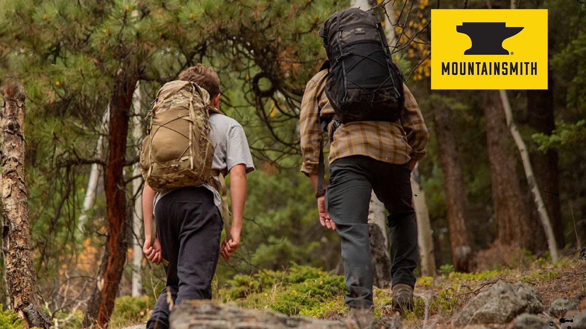 Two hikers wearing Mountainsmith backpacks walk away on a forest trail surrounded by evergreen trees and moss-covered rocks, showcasing outdoor gear for backcountry adventure.
