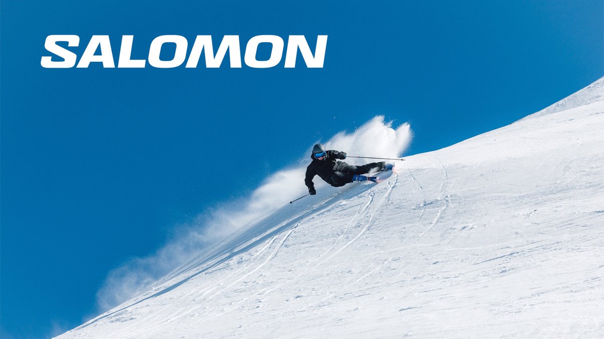 A skier in all-black gear carves a sharp turn down a steep, sunlit snow slope under a clear blue sky.