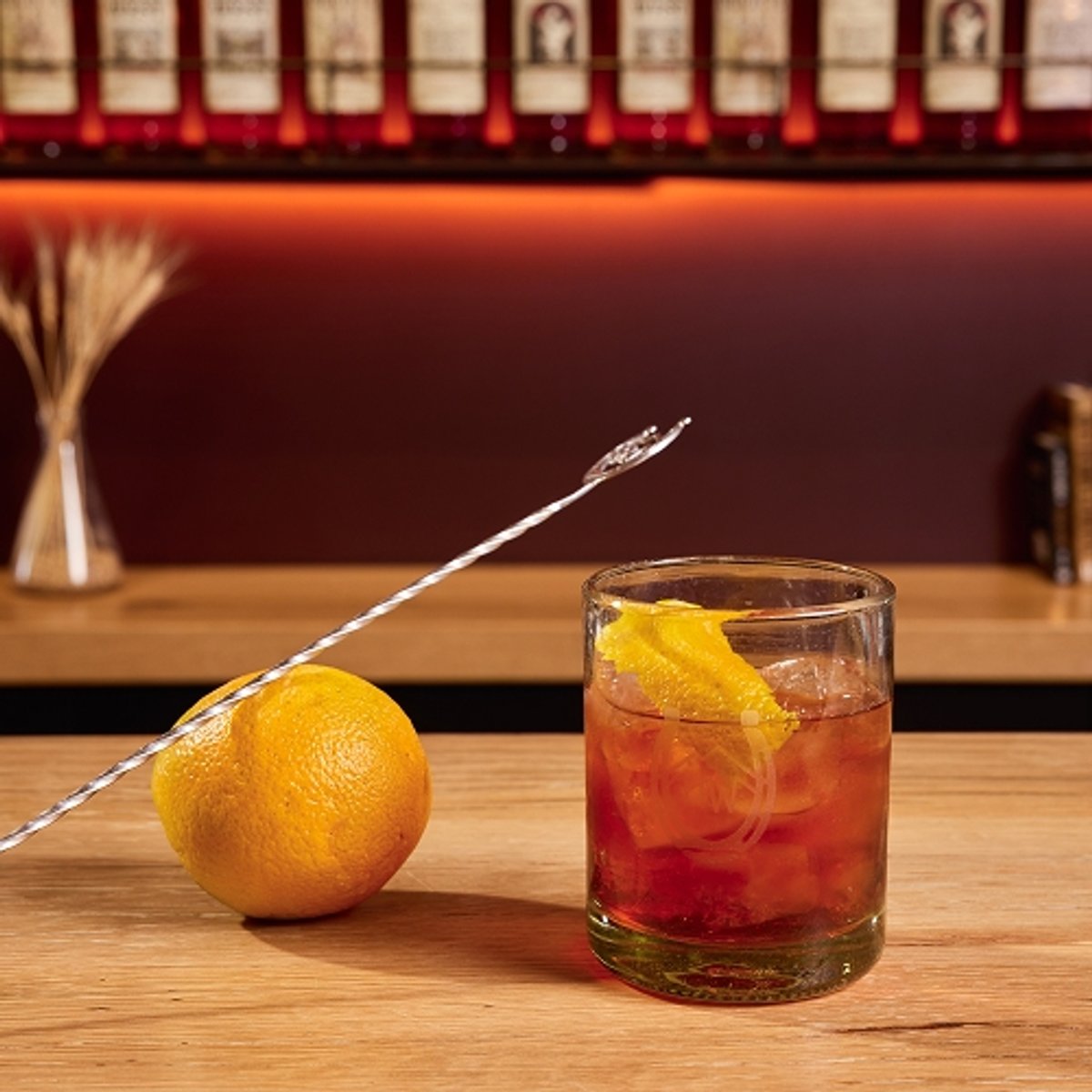 A bar countertop displays ingredients for a Strawberry Boulevardier cocktail: a glass with an orange twist and red drink, an orange, a long stirring spoon, a bottle of High West Bourbon, a bottle of sweet vermouth, and a bottle of Campari, with a background of liquor bottles and large text reading "Strawberry Boulevardier" below the High West Distillery and Backcountry logos.