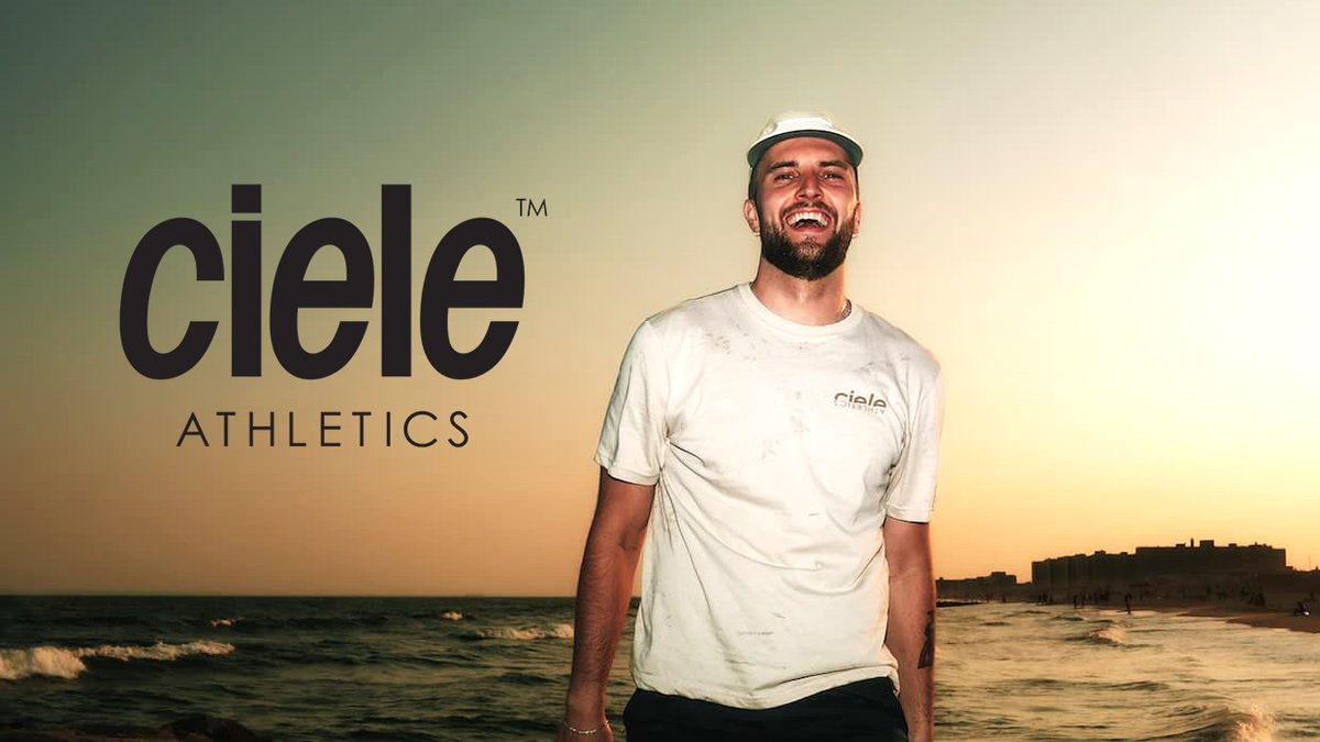 Man wearing white Ciele Athletics t-shirt and cap standing on beach at sunset with ocean waves and coastal buildings in background.
