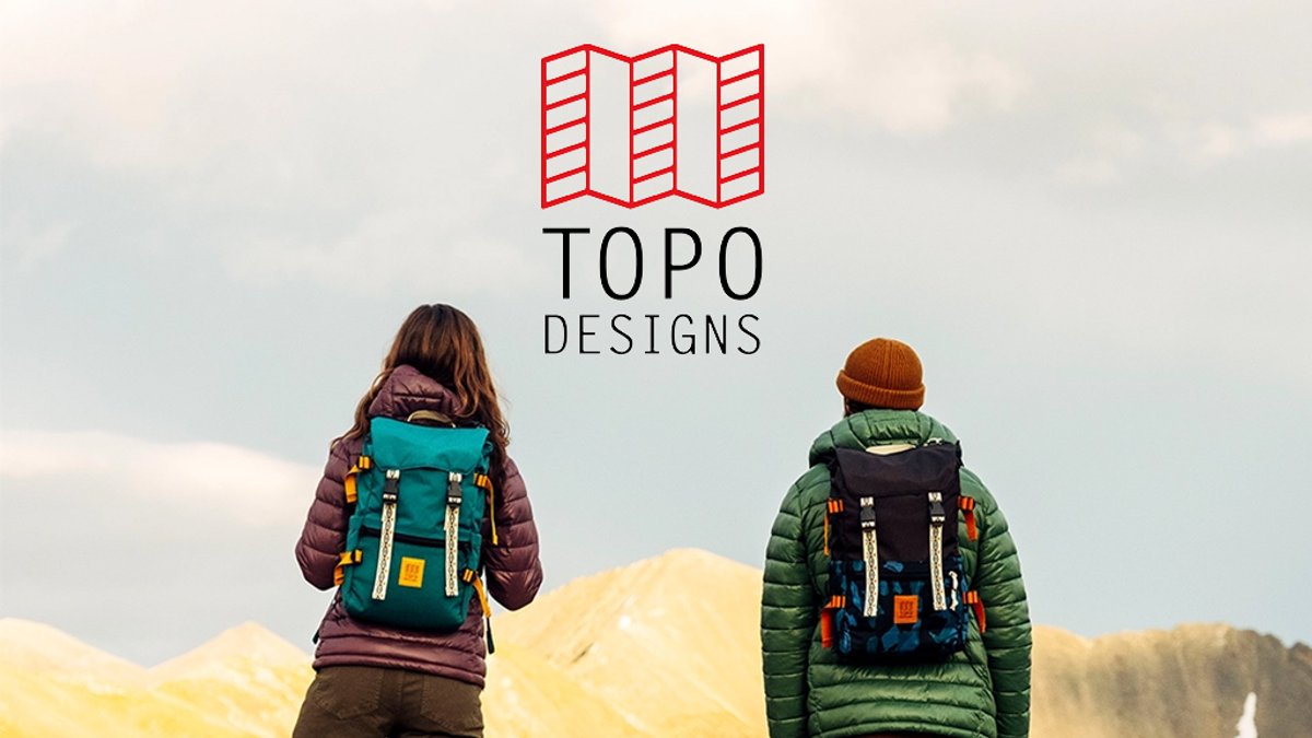 Two hikers wearing colorful backpacks and winter jackets stand facing away from the camera overlooking a mountainous landscape beneath the Topo Designs logo.