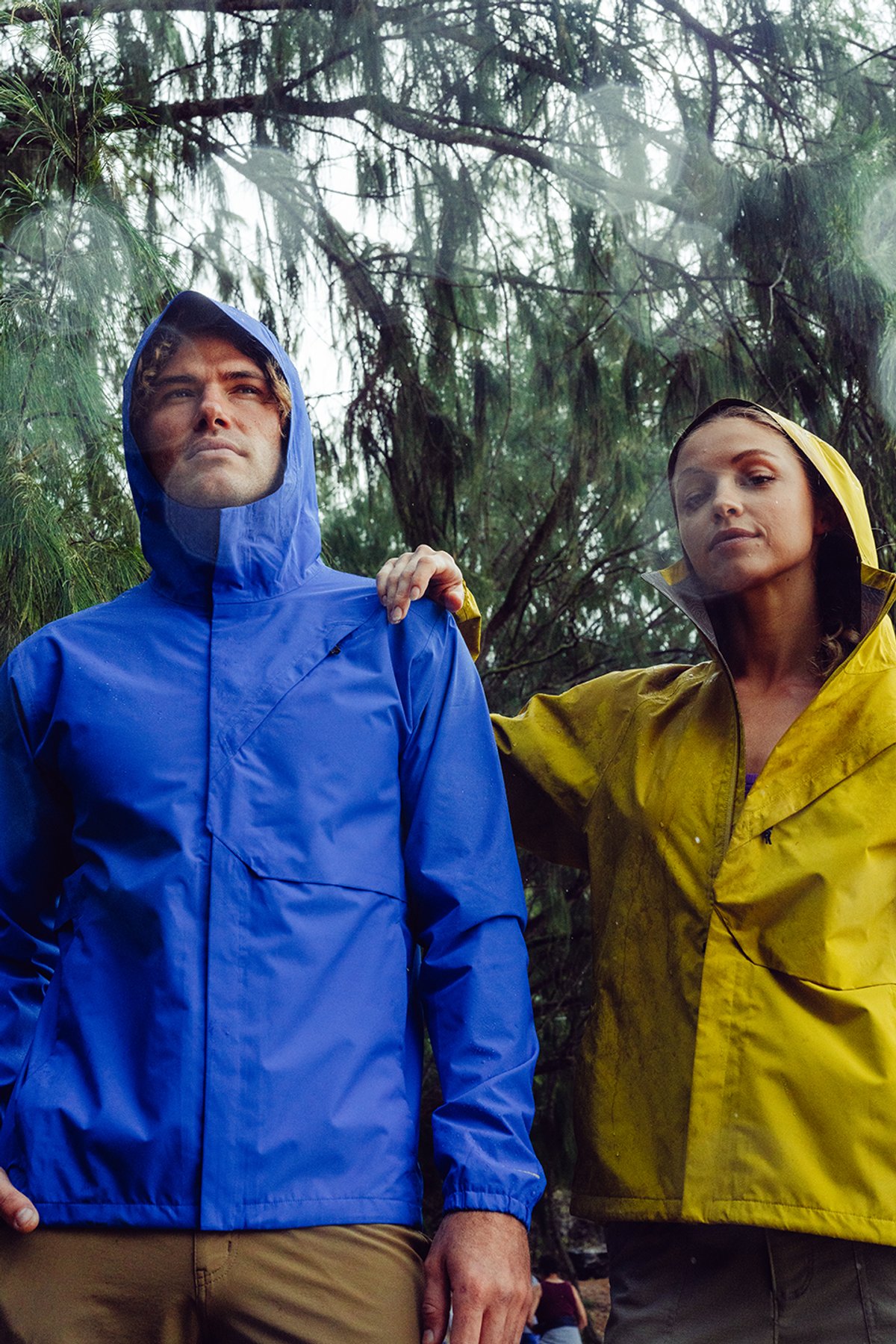 Man and woman wearing blue and yellow hooded waterproof rain jackets standing in wet tropical forest during rain