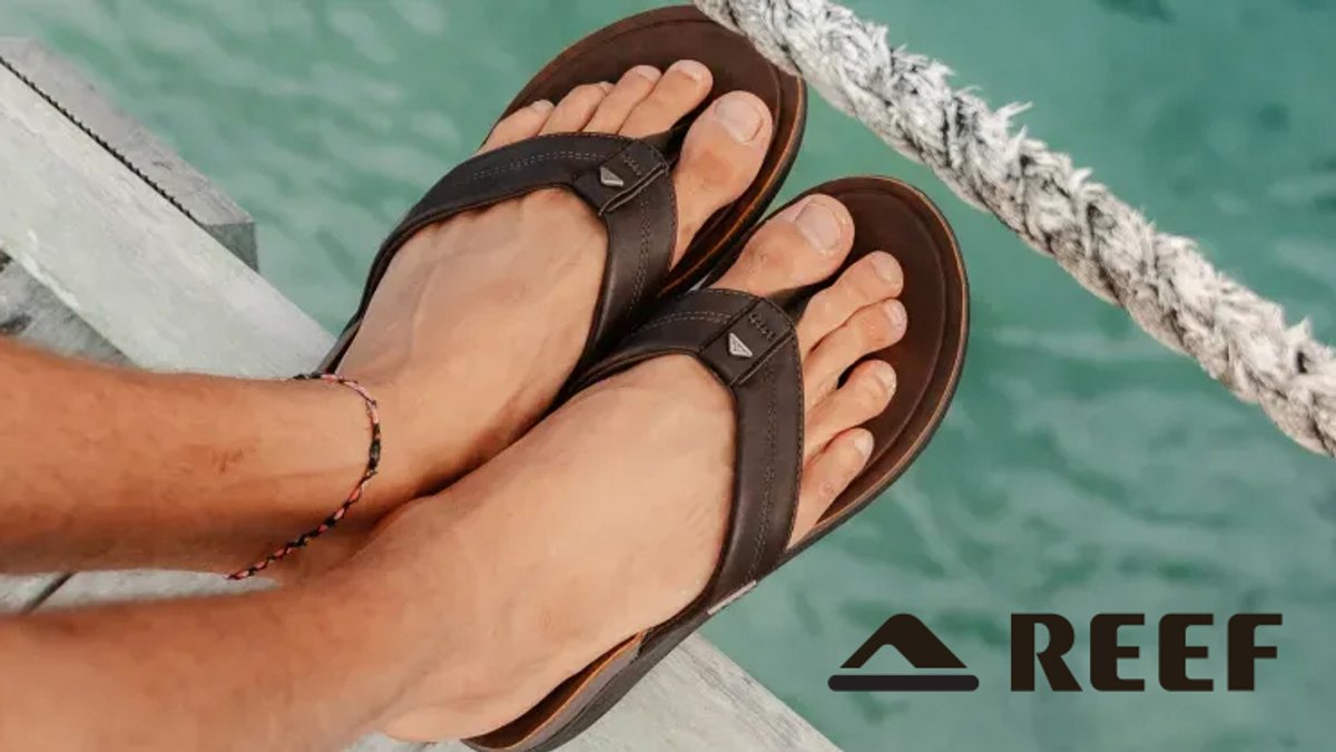 Person wearing black Reef sandals with brown soles sitting on a white dock over turquoise water.
