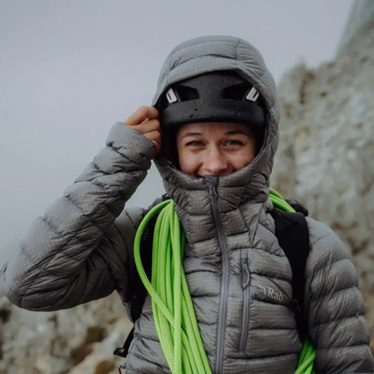 A woman wears a Rab Microlight jacket with the hood over her climbing helmet.