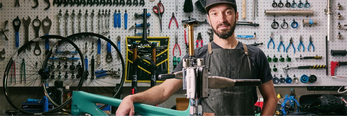 A man in a bike shop stands over a bike frame. He’s looking at the camera with a face that says, “I can build any bike, try me.” 