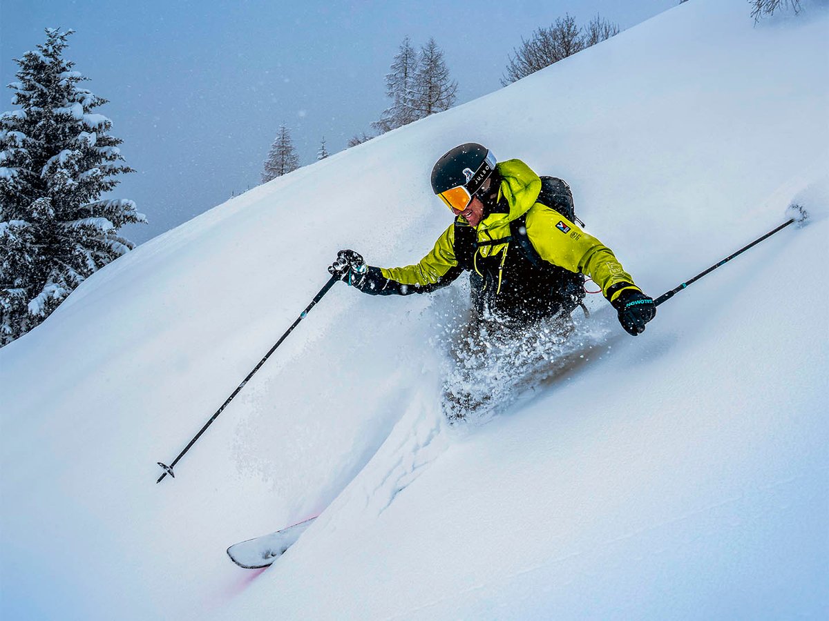 Backcountry skier charging through deep powder on steep terrain, neon jacket, snow-covered trees in background
