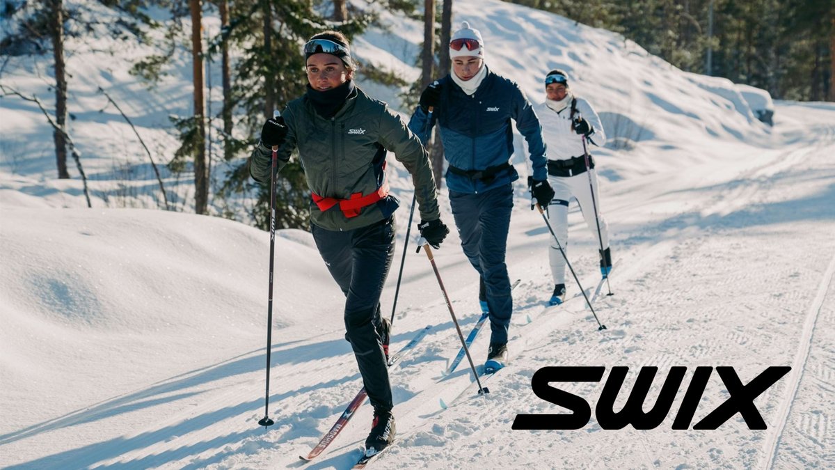 Three cross-country skiers in Swix winter apparel skiing on a snowy mountain trail surrounded by evergreen trees on a sunny day.